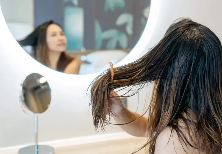 Una joven se peina el pelo mojado frente a un espejo. El pelo adolescentes, largo y castaño, cae en mechones.