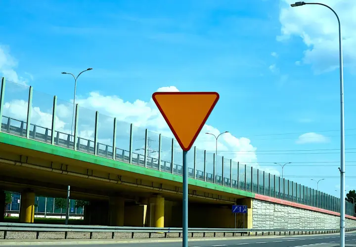Pomarańczowy znak drogowy "ustąp pierwszeństwa" na tle wiaduktu i błękitnego nieba.