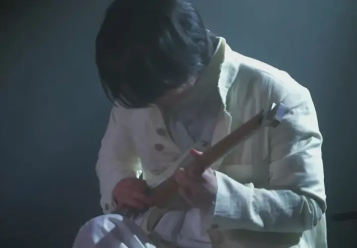 Ein Musiker in weißer Kleidung spielt eine Gitarre. Die Szene ist düster beleuchtet, was die Konzentration auf die Performance von Ichika Nito verstärkt.