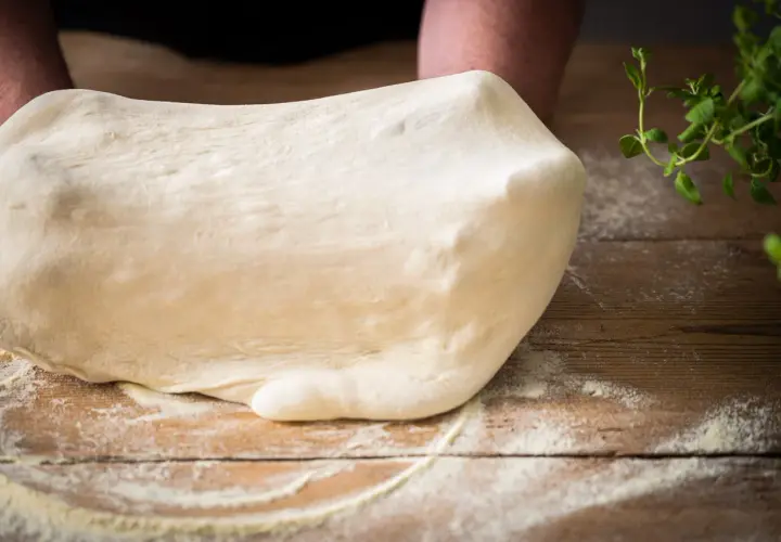 Frischer Teig für Pizza Semola wird auf einem Holztisch geknetet, bestäubt mit Mehl. Kräuter liegen daneben.