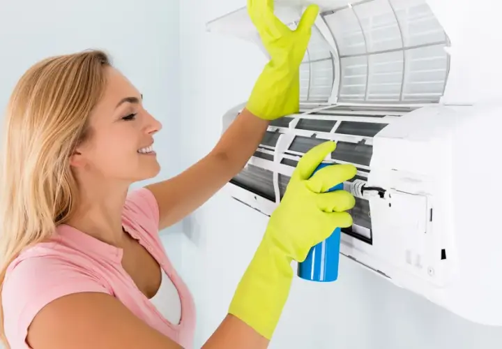 Mujer con guantes verdes limpia el aire acondicionado que huele a vinagre, rociando un producto para eliminar olores.