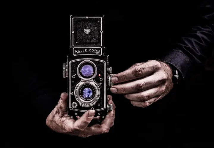 Dwie dłonie trzymają zabytkowy aparat Rolleiflex, którego obiektywy odbijają światło, tworząc wrażenie złotych proporcji w kadrze.