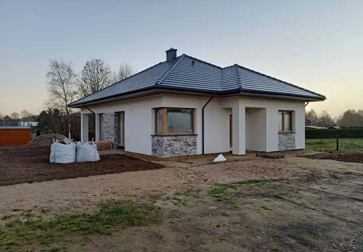 Nowy, nowoczesny dom w renklodach 15 z szarym dachem i białymi ścianami, otoczony placem budowy.
