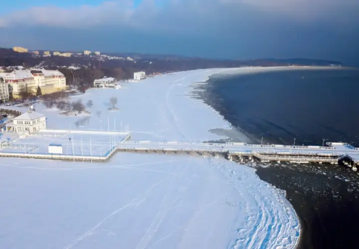 Zimowy spacer po molo, gdzie zimą nad morze przyciąga biel śniegu i lodowe krajobrazy. Widok na zamarznięte plaże i budynki w oddali.