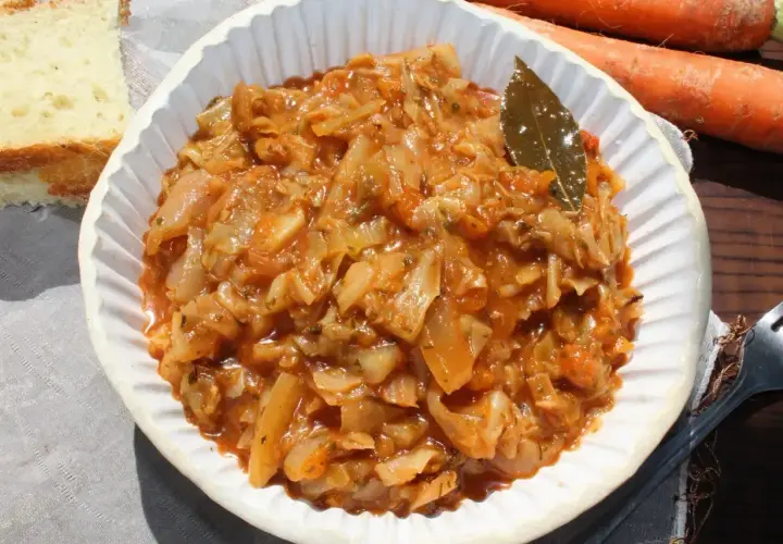 Pyszny bigos z młodej kapusty w białej misce, obok kromka chleba i marchewki.
