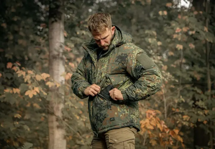 Mężczyzna w kurtce w kamuflaże wojskowe, zapinanej na zamek, stoi w lesie.