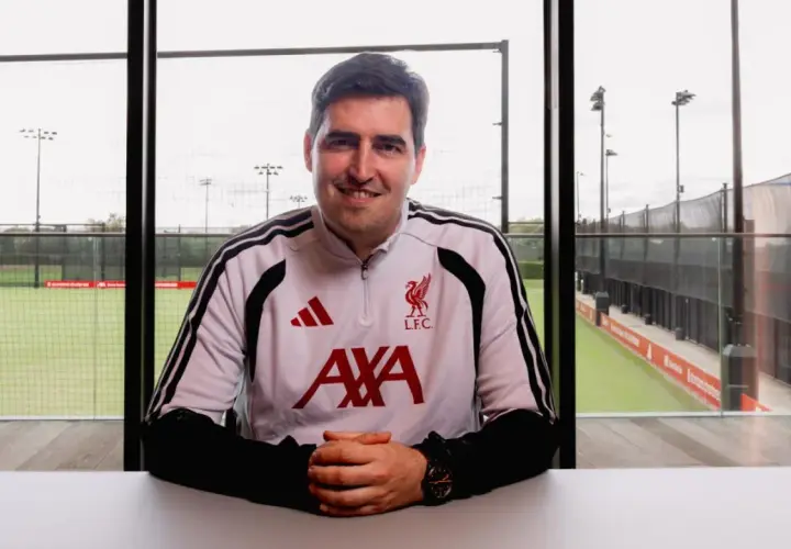 Uśmiechnięty trener Liverpoolu w stroju treningowym z herbem klubu i logo AXA. Za nim boisko treningowe.