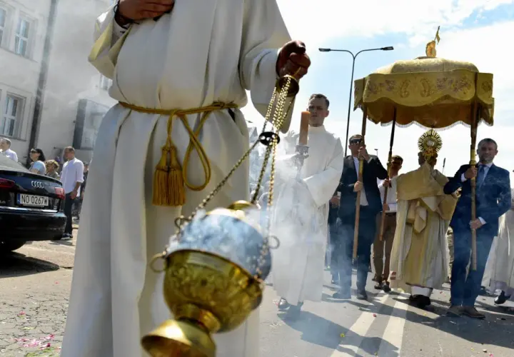 Procesja Bożego Ciała z kadzidłem i monstrancją. Uczestnicy w białych szatach idą ulicą.
