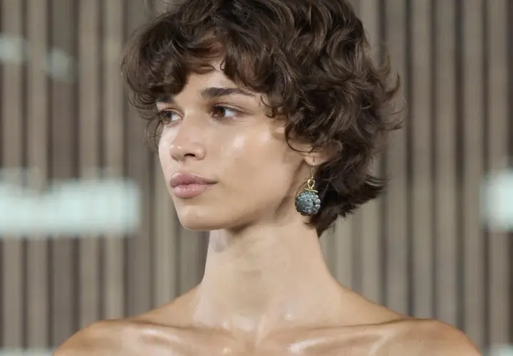 Mujer con pelo corto fino y escaso, rizado y castaño. Luce un pendiente colgante con una piedra grisácea.