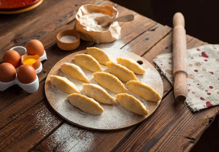 Surowe pierogi ułożone na desce, obok jajka, mąka i wałek.