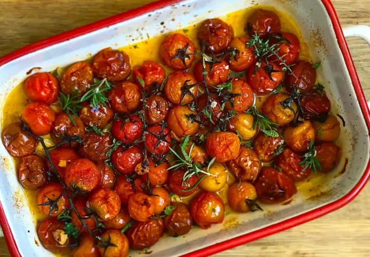 Tomates cherry confitados en aceite de oliva, con romero y tomillo, en una bandeja blanca con borde rojo.