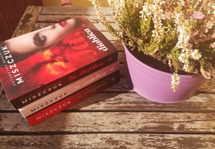 Na drewnianym stole leżą trzy książki Katarzyny Miszczuk: "Ja, diablica", "Ja, anielica" i "Ja, potępiona". Obok stoi fioletowa doniczka z wrzosem. Wszyscy jesteśmy w grze.