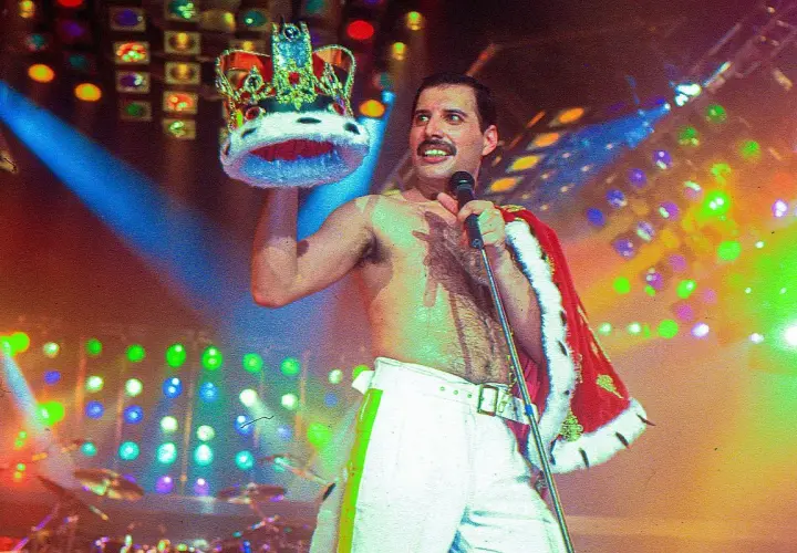 Freddie Mercury, w koronie i z mikrofonem, na scenie pełnej świateł. Pamięć o nim żyje, choć zmarł Freddie Mercury.