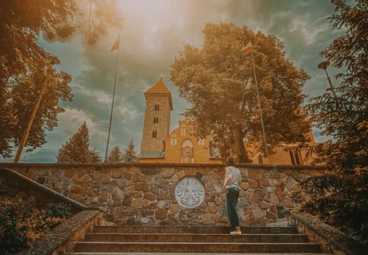Osoba na schodach przed kamiennym murem z godłem Polski, w tle kościół w Czerwińsku nad Wisłą.