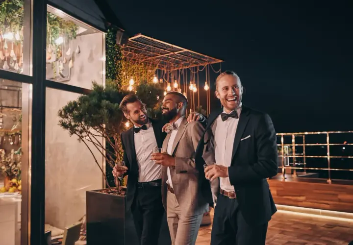Trzech mężczyzn w eleganckich garniturach, z drinkami w rękach, śmieje się i rozmawia na tarasie. Jeden z nich, jako drużba, opiera rękę na ramieniu przyjaciela.