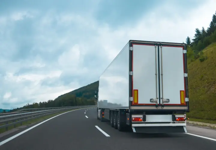 Ciężarówka na autostradzie, z tyłu widać białą naczepę. Na poboczu rosną drzewa, a nad nimi chmury. Tablice wyróżniające przepisy są widoczne na naczepie.
