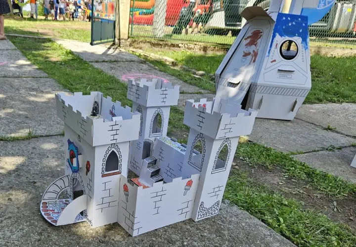 Zabawki z kartonu diy: biały zamek z wieżami i schodami, obok rakieta. Dzieci bawią się w tle.