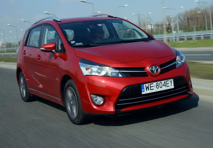 Czerwona Toyota Corolla Verso jedzie po drodze. Kierowca w okularach przeciwsłonecznych prowadzi auto.