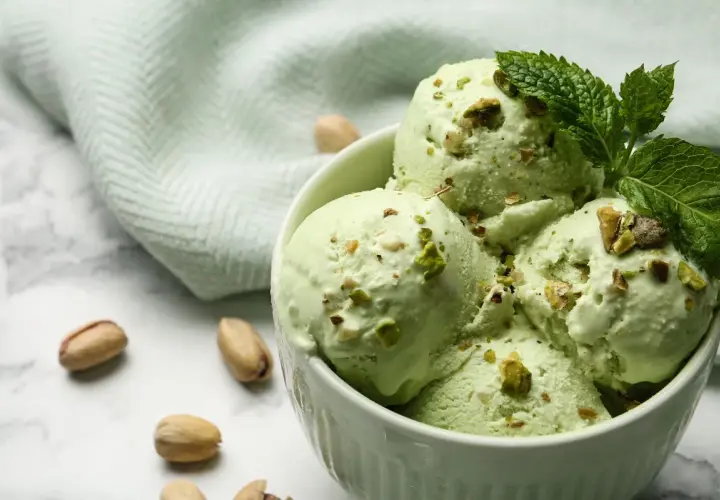Coppa di cremoso gelato al pistacchio, guarnita con foglioline di menta e granella di pistacchi. Alcuni pistacchi interi sparsi sul marmo.