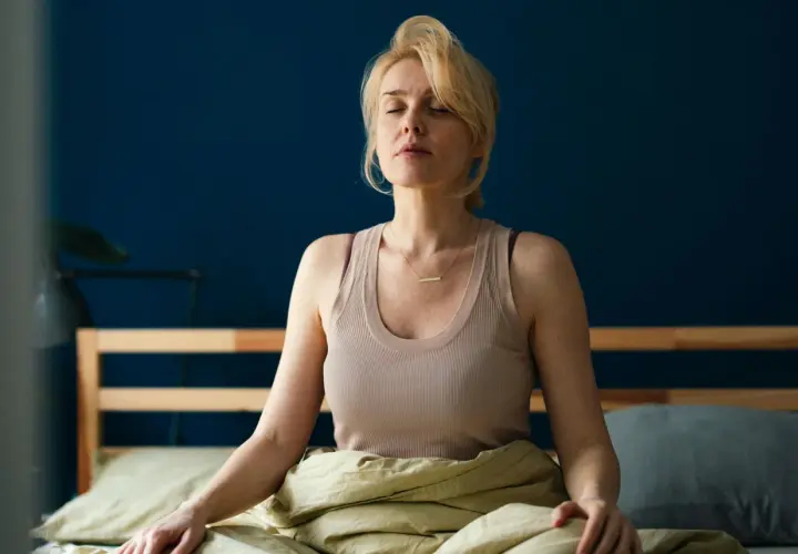 Kvinna med slutna ögon sitter i sängen, insvept i täcke, i en avslappnad somna meditation.