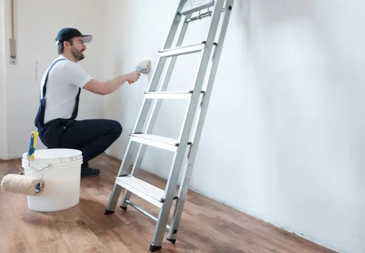 Hombre con brocha y cubo de pintura, listo para limpiar pared antes de pintar. Escalera metálica al lado.