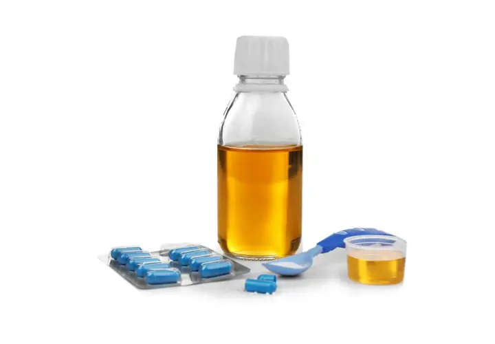 Flacone di sciroppo amoxicillina bambini, blister di capsule blu e misurino dosatore con polvere bianca.