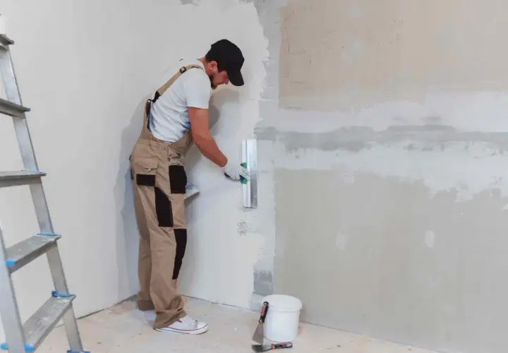 Operaio con tuta beige e cappellino nero mentre impara come intonacare un muro, usando una spatola. Scala e secchio a terra.