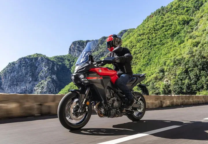 Un motociclista su una Yamaha Tracer rossa affronta una curva su una strada panoramica, godendosi una gita in moto tra montagne verdi e cielo azzurro.