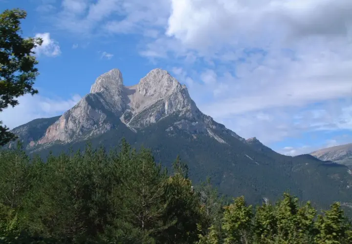 Majestuoso Pedraforca se alza imponente, su altitud desafía las nubes. Bosques verdes cubren sus faldas bajo un cielo azul.