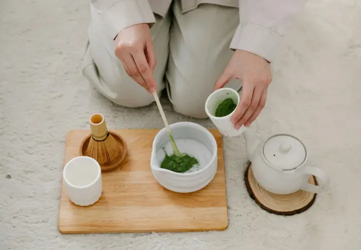 Hände bereiten Matcha zu. Ein Bambusbesen und eine Tasse stehen bereit. Dieser Matcha macht wach!