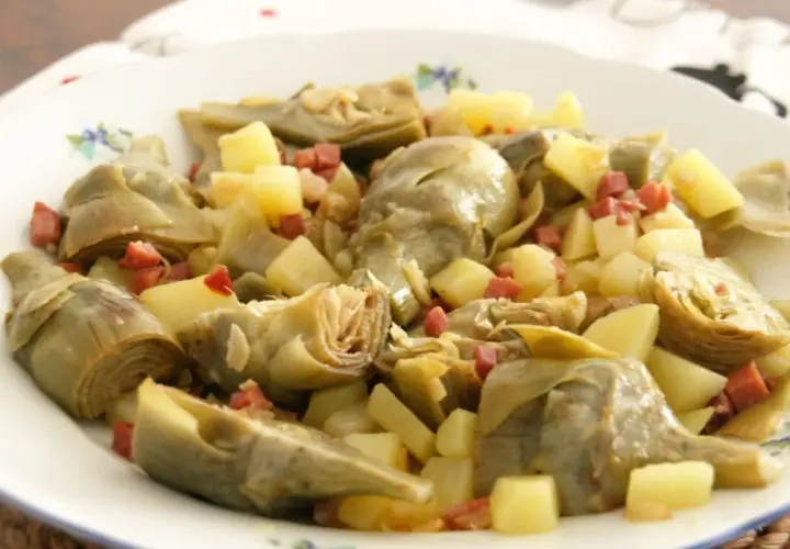 Plato de alcachofas tiernas y patatas cortadas en cubos, salpicado con trocitos de jamón. Un manjar casero.