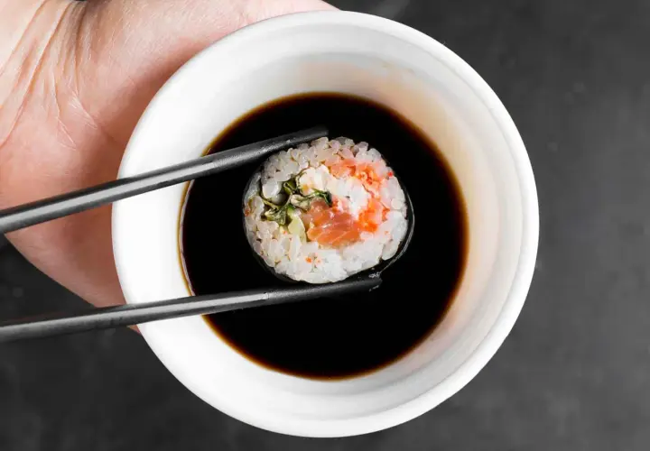 Pałeczkami zanurzam rolkę sushi z łososiem w ciemnym sosie.