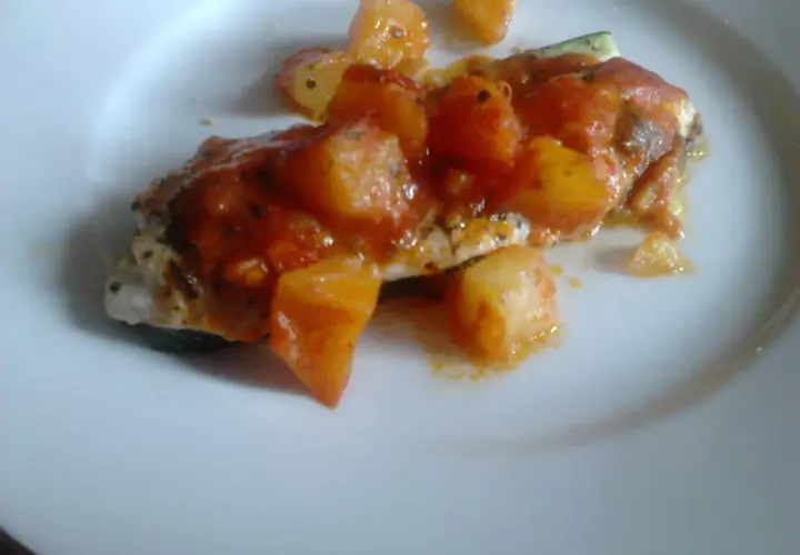 Un delicioso jargo pescado cubierto con salsa de tomate y trozos de patata, servido en un plato blanco.