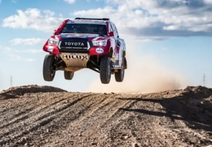 Toyota Hilux w locie podczas rajdu Dakar. Widzowie obserwują zmagania na pustynnym terenie.