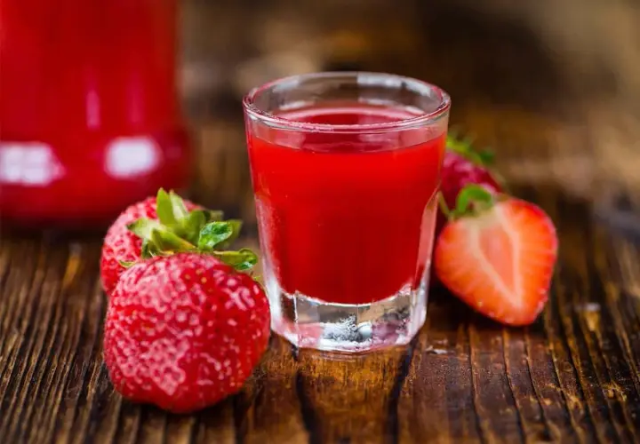 A shot glass filled with vibrant red nalewka z truskawek, surrounded by fresh strawberries on a wooden surface.