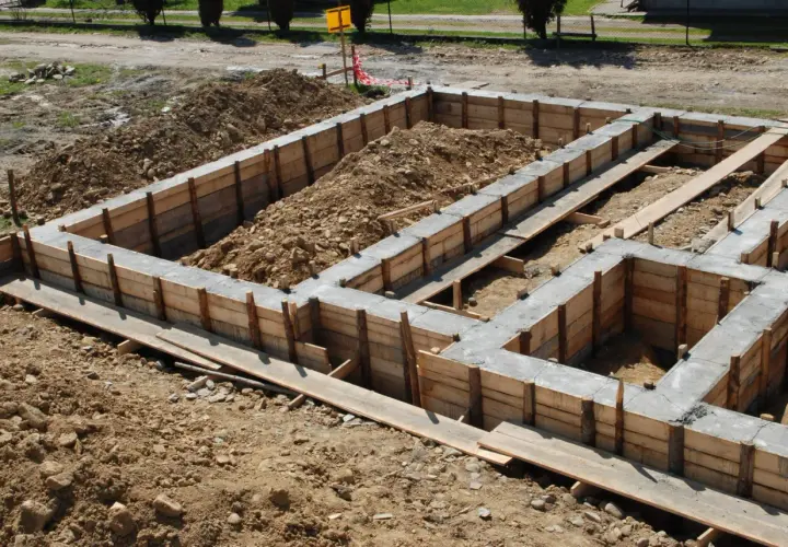 Budowa domu w toku: betonowe ławy fundamentowe otoczone drewnianym szalunkiem, zasypane ziemią.