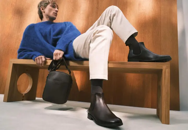 Mężczyzna w niebieskim swetrze i beżowych chinosach prezentuje stylowe buty do chinosów. Elegancka torba na ramię dopełnia całość.