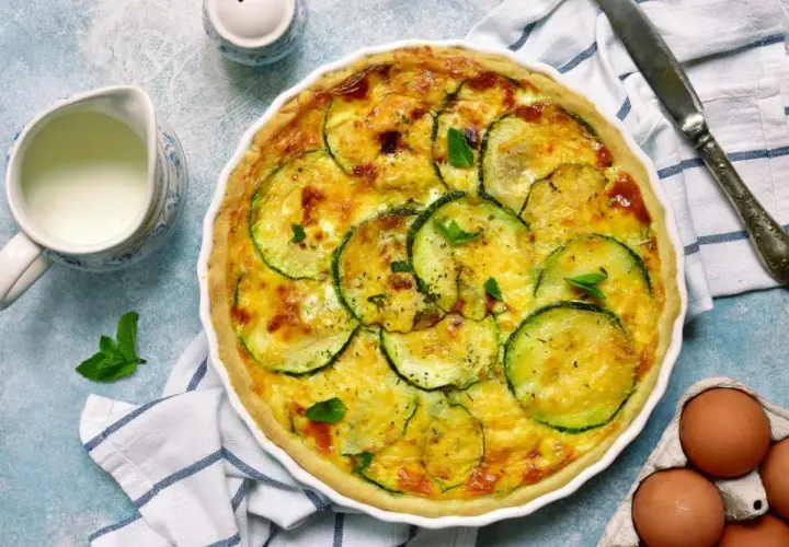 Pyszna tarta z cukinią, zapieczona ze złotym serem i ziołami. Obok dzbanuszek mleka i jajka.