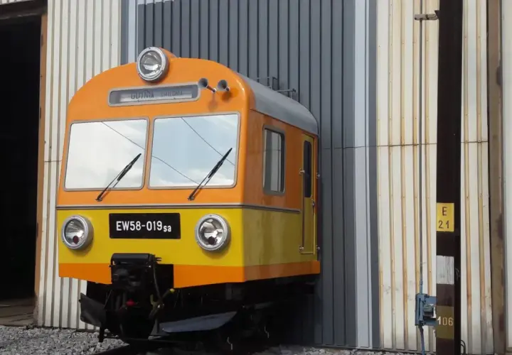 Pomarańczowo-żółty pociąg EW58-019sa stoi przed halą.
