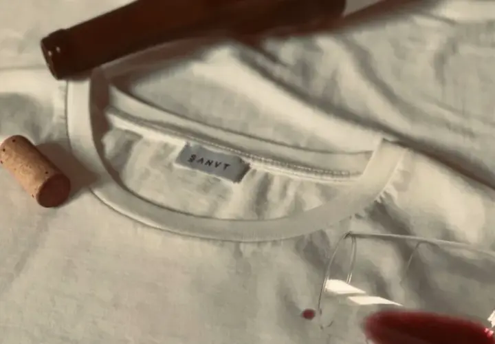Un bouchon de liège et une bouteille de vin à côté d'un tee shirt blanc, avec une tache de vin rouge.
