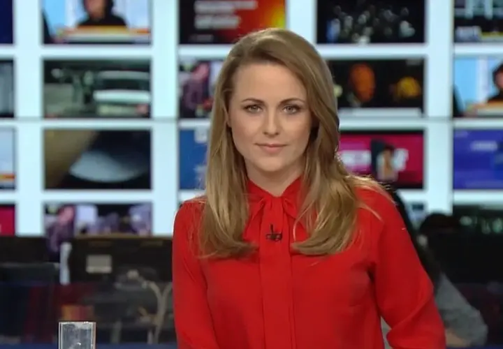 Marta Kłos, której wiek nie jest widoczny, w czerwonej bluzce z kokardą, na tle ekranów informacyjnych.