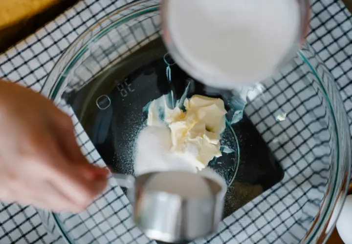 Kobieta wsypuje cukier do miski z masłem na wadze kuchennej.