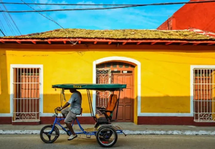 Un hombre pedalea un taxi bicitaxi frente a una casa amarilla vibrante, un recuerdo de mi viaje a Cuba.
