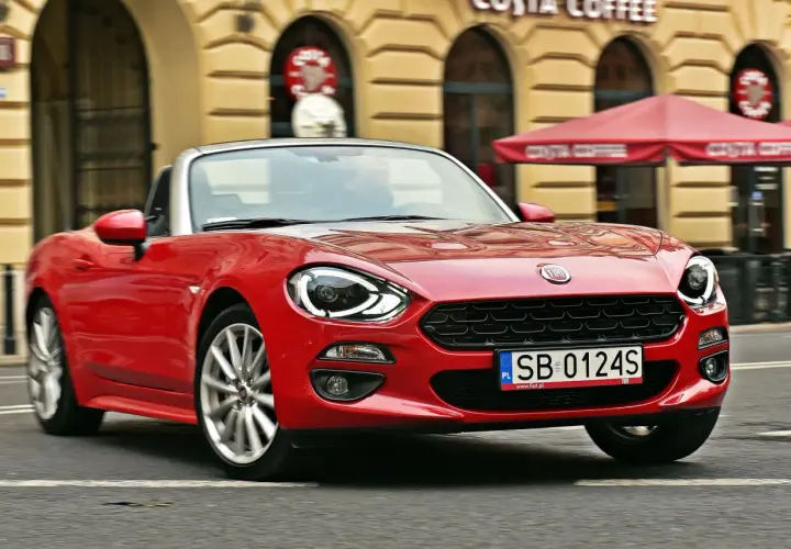 Czerwony Fiat 124 Spider z otwartym dachem sunie ulicą przed kawiarnią Costa Coffee.