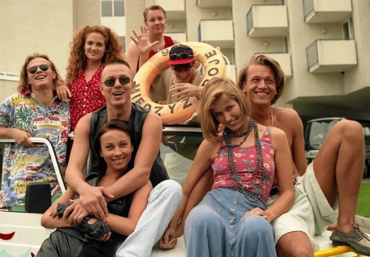 Grupa uśmiechniętych polscy aktorzy lat 90. pozuje na tle hotelu, niektórzy siedzą na samochodzie, inni stoją.