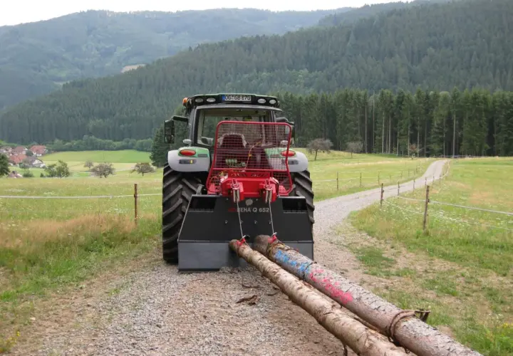Traktor mit angehängter seilwinde forst zieht zwei Baumstämme über einen Schotterweg, umgeben von grünen Wiesen und einem dichten Wald.