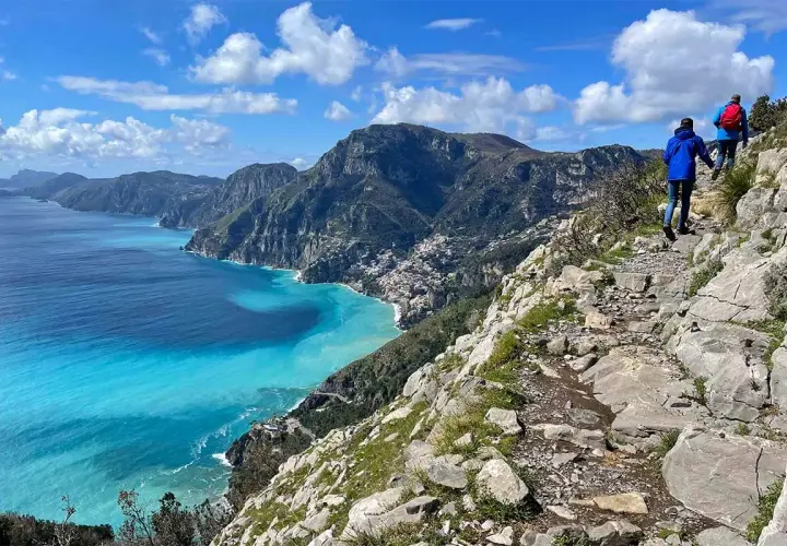 Hikers on the "walk of the gods" trail, with stunning turquoise waters and coastal villages below.
