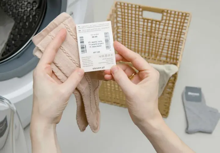 Dłonie trzymające beżowe skarpetki z metką obok koszyka i pralki.