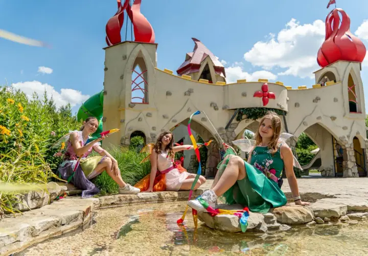 Trzy dziewczynki w strojach wróżek siedzą nad wodą w bajkowym parku rozrywki dla dzieci.