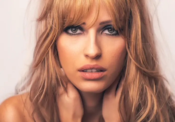 Kobieta z blond włosami i cieniowaniem grzywki, z dłońmi na szyi.